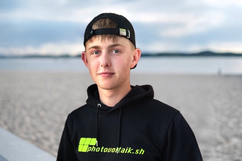 Junger Mann mit schwarzem Kapuzenpulli und umgekehrter Mütze steht an einem Sandstrand mit Wasser und entferntem Land im Hintergrund.