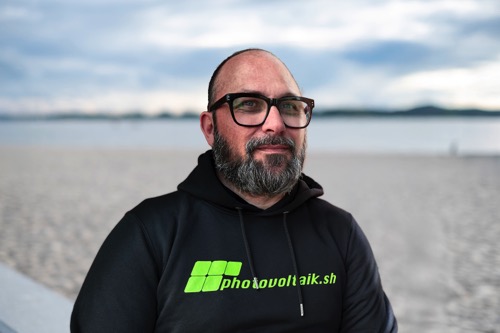 Ein Mann mit Brille und Bart, der einen schwarzen Kapuzenpulli mit dem Aufdruck "photovoltaik.sh" trägt, sitzt an einem Strand mit einem bewölkten Himmel im Hintergrund.