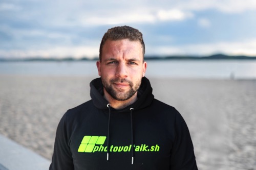 Ein Mann mit Bart und einem schwarzen Kapuzenpulli mit grüner Aufschrift steht an einem Sandstrand mit einem ruhigen Gewässer im Hintergrund.